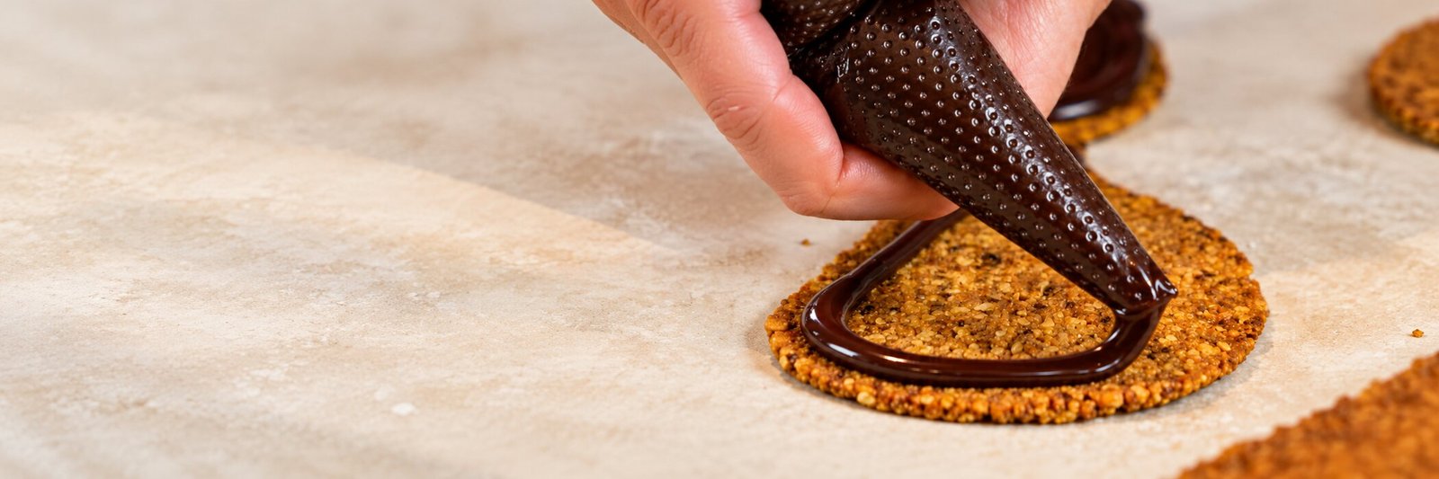 Une main qui poche du chocolat fondu sur un biscuit aux fruits secs, sur un plan de travail fariné